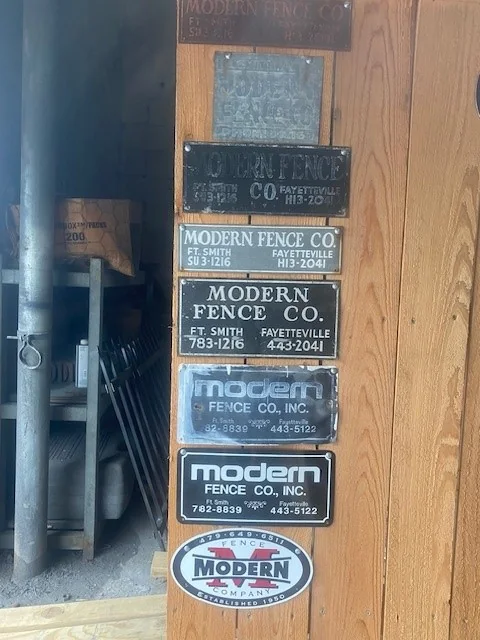 Modern Fence Company nameplates from several decades mounted one above another, from cast metal plates to the current logo